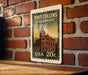 A metal sign with a vintage-style illustration of a building and a clock, mounted on a brick wall.