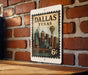 A metal sign with a vintage-style illustration of the Dallas skyline, mounted on a brick wall.
