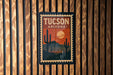 A vintage-style postage stamp depicting the city of Tucson, Arizona, with a cactus landscape and a sunset in the background.