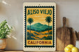 a vintage-style sign for Aliso Viejo, California, with a palm tree and landscape illustration. The sign is displayed on a shelf next to a wooden cutting board and a potted plant.