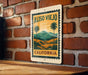 A vintage-style metal sign with the text "ALISO VIERO CALIFORNIA" and a landscape illustration of a tropical setting, mounted on a brick wall.