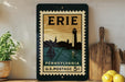 A framed vintage postage stamp from Pennsylvania, featuring a lighthouse and a sailboat, is displayed on a shelf next to a wooden cutting board and a potted plant.