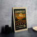 A vintage postage stamp from California, featuring a landscape with mountains, trees, and a sunset, placed on a countertop next to some black camera lenses.