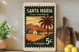 A vintage-style postage stamp from Santa Maria, California, featuring a rural landscape with a palm tree and a house, displayed on a shelf next to a wooden cutting board and some lemons.