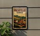 A vintage-style postage stamp depicting a cityscape with mountains in the background, mounted on a wall next to a plant.