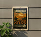 A postage stamp-like sign with a scenic landscape of mountains, fields, and a sunset, mounted on a wall.