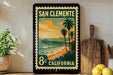 A framed vintage postage stamp depicting a beach scene with a pier and palm trees, placed on a shelf next to a wooden cutting board and a potted plant.