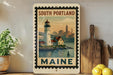 A framed vintage-style poster of a lighthouse and a cityscape in Maine, with the text "SOUTH PORTLAND MAINE" prominently displayed.