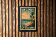 A vintage-style postage stamp from San Clemente, California, featuring a beach scene with palm trees and a pier.