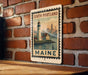 A vintage-style metal sign depicting a lighthouse and a cityscape, with the text "SOUTH PORTLAND MAINE" printed on it, is mounted on a brick wall.