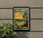 A vintage-style postage stamp depicting a beach scene with palm trees and a pier, mounted on a wall.