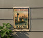 A framed poster of a lighthouse and a cityscape, with the text "SOUTH PORTLAND" and "MAINE" printed on it, is displayed on a wall next to a plant.