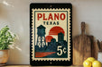 A vintage-style postage stamp from Plano, Texas, featuring a water tower and sunset, displayed on a shelf with a wooden cutting board and a potted plant.