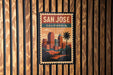 A vintage-style poster of San Jose, California, featuring a cityscape with palm trees and mountains, mounted on a wooden wall.