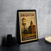 A framed poster of Brighton, Colorado, featuring a water tower and a building, is displayed on a countertop.