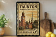 A framed vintage postage stamp from Massachusetts, depicting a church and other buildings, is displayed on a shelf next to a wooden cutting board and some lemons.