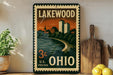 A vintage-style metal sign with the text "LAKEWOOD OHIO 3c U.S. POSTAGE" displayed on a shelf, with a potted plant and wooden cutting board in the background.