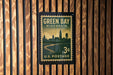 A vintage-style postage stamp from Wisconsin, featuring a silhouette of a city skyline and a river, mounted on a wooden wall.