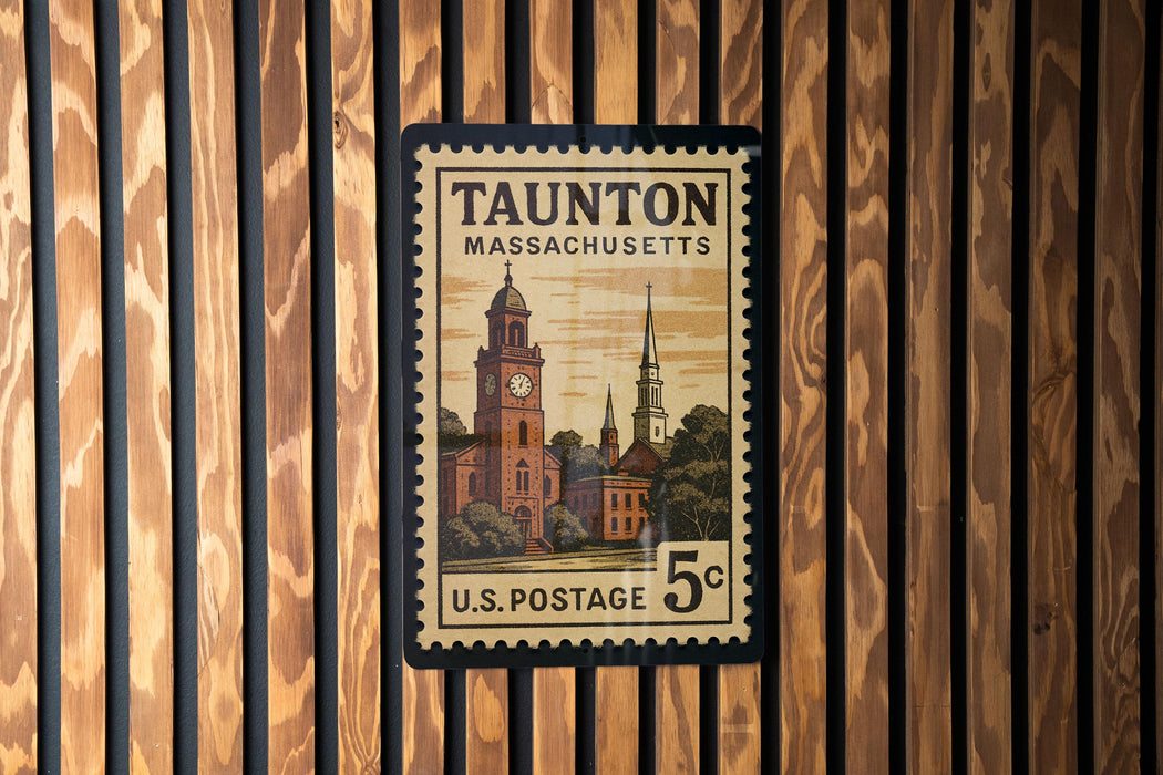 A framed U.S. postage stamp from Massachusetts, featuring a clock tower and other architectural elements, is displayed on a wooden wall.