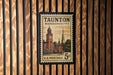 A framed U.S. postage stamp from Massachusetts, featuring a clock tower and other architectural elements, is displayed on a wooden wall.