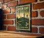 A vintage-style postage stamp from Wisconsin, featuring a cityscape and a river, is displayed on a brick wall.