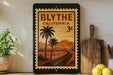 a framed poster of a tropical road scene with palm trees and mountains, placed on a shelf next to a wooden cutting board and some lemons.