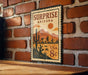 A vintage-style metal sign with a desert landscape and mountains, mounted on a brick wall.