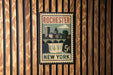 A vintage postage stamp from Rochester, New York, featuring a cityscape and a waterfall, mounted on a wooden wall.