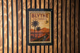 A vintage postage stamp from California, featuring a palm tree and a road, is displayed on a wooden wall.