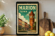 A framed vintage-style poster of Marion, Iowa, featuring a clock tower and a row of colorful buildings, is displayed on a shelf next to a wooden cutting board and some lemons.
