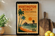 A framed poster of Huntington Beach, California, featuring a tropical beach scene with palm trees, a pier, and a sunset.