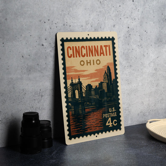 A vintage-style postage stamp from the United States, featuring an illustration of the Cincinnati skyline and a bridge, placed on a gray surface next to some black camera lenses.
