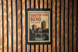 A vintage-style poster of South Bend, Indiana, featuring a cityscape and a bridge, is displayed on a wooden wall.