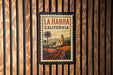 A framed poster of La Habra, California, featuring a palm tree and a road leading to a church, is displayed on a wooden wall.