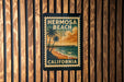 A vintage-style poster of Hermosa Beach, California, featuring a beach scene with palm trees and a pier, is displayed on a wooden wall.