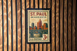 A postage stamp from Minnesota, featuring a cityscape and a bridge, is displayed on a wooden wall.
