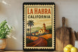 A framed poster of La Habra, California, featuring a scenic illustration of a road leading to a church, surrounded by palm trees and hills.