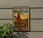 A vintage-style metal sign with a colorful illustration of a town, mounted on a wall next to a plant.