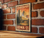 A vintage-style postage stamp from Iowa featuring a cityscape with a bridge and trees, mounted on a brick wall.