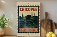 A framed vintage-style stamp depicting the city of Chicopee, Massachusetts, with a bridge and buildings in the background.