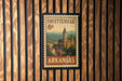 A vintage-style postage stamp from Arkansas, featuring a building and landscape, is displayed on a wooden wall.