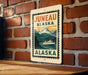 A metal sign with the text "JUNEAU ALASKA" and an illustration of a boat on a river, mounted on a brick wall.