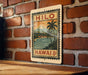 A vintage-style metal sign with a tropical landscape and palm trees, featuring the text "HILO" and "HAWAII 8¢", is displayed on a brick wall.