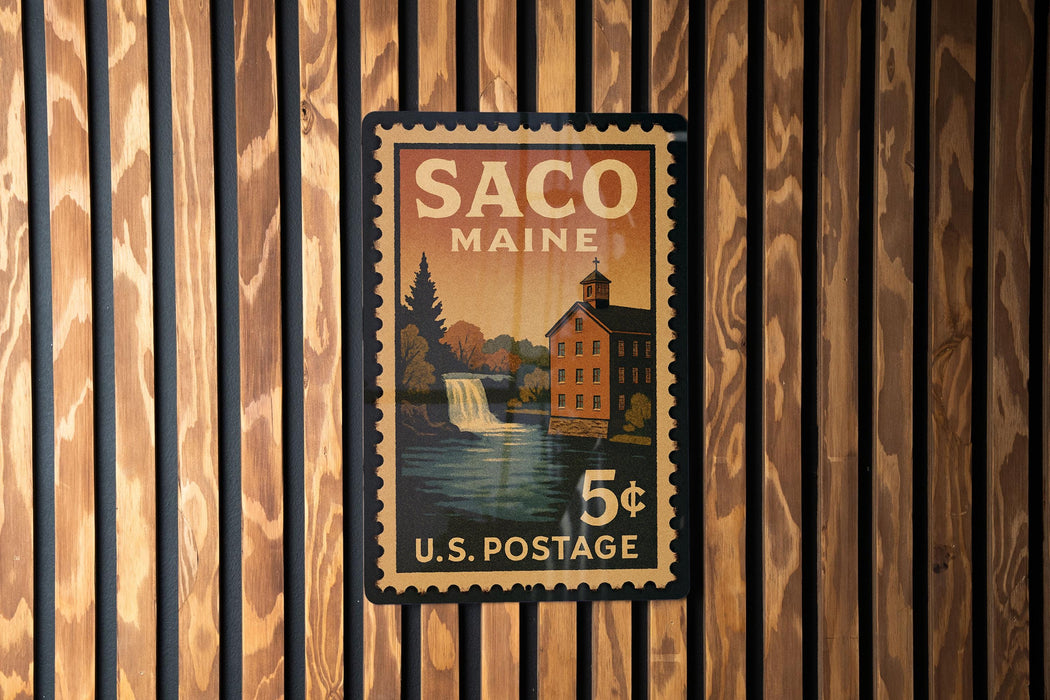 A vintage U.S. postage stamp from Maine, featuring a waterfall and a building, is displayed on a wooden wall.