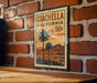 A vintage-style metal sign with a palm tree and sunset landscape, mounted on a brick wall.