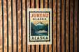a sign that reads "Juneau Alaska" with a mountain landscape and a boat in the background.