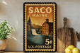 A vintage postage stamp from Maine, featuring a scenic illustration of a waterfall and a building, displayed on a shelf next to a wooden cutting board and some lemons.