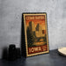 A vintage-style postage stamp from Cedar Rapids, Iowa, featuring a cityscape with a bridge and trees, placed on a countertop next to some black camera lenses.
