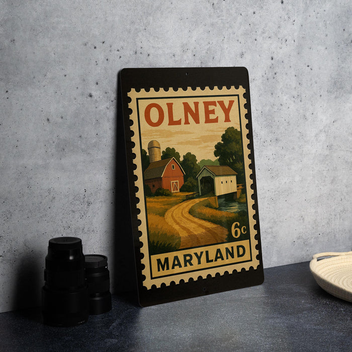 A metal stamp depicting a rural scene with a barn, farmhouse, and road, placed on a countertop next to some black camera lenses.