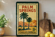 A vintage-style sign for Palm Springs, California, featuring palm trees and mountains, is displayed on a shelf next to a wooden cutting board and some lemons.